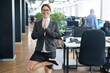 © Михаил Решетников - A pacified business woman doing yoga in an open space office. A red-haired smiling female employee in a casual suit and glasses stands in a tree pose at the workplace. Meditation and relaxation.