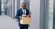 © VAKSMANV - Sad young African American man in suit and tie walking the street, carrying box with stuff after fired from work. Jobless male office worker strolling outdoor. Fire during lockdown crisis.