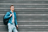 Fototapeta Na drzwi - Young handsome man in shirt, jeans and glasses carrying a backpack and holds smartphone while standing on a background of a gray wall. Copy, empty space for text