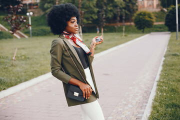 Wall Mural - Fashionable girl in a summer city. Black lady in a green jacket. Woman in a red scarf. Famale with mobile phone