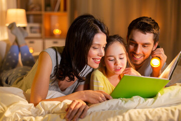 Wall Mural - people and family concept - happy mother, father and little daughter reading book with torch light in bed at night at home