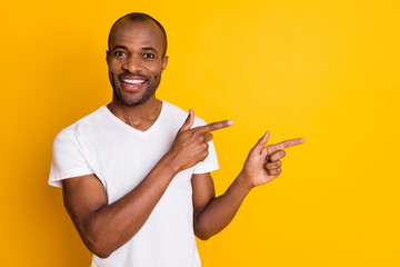 Wall Mural - Photo of attractive dark skin guy hold arms directing fingers side empty space showing novelty information wear casual white t-shirt isolated bright vivid yellow color background