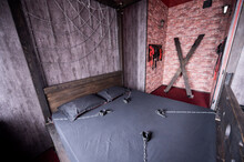 Torture Of Andreev Cross On The Bedroom Wall. VDSM Equipment And Sex Toys. Leather Handcuffs With A Chain On A Gray Sheet. The Lashes Hang In The Room For Perversions.