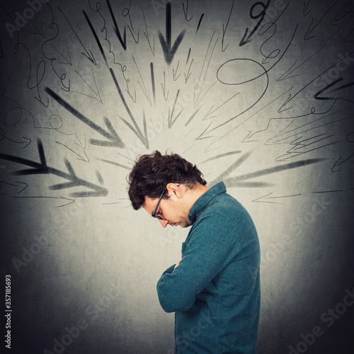 Upset introvert person looking down troubled, feels dismayed. Lonely guy experiencing emotional crisis. Man being under pressure at workplace as multiple arrows on the wall pointing to him.