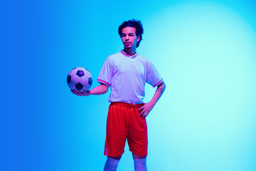 Wall Mural - Leader. Football or soccer player on gradient blue studio background in neon light - posing confident with ball. Concept of sport, competition, winning, action, motion, overcoming. Copyspace.