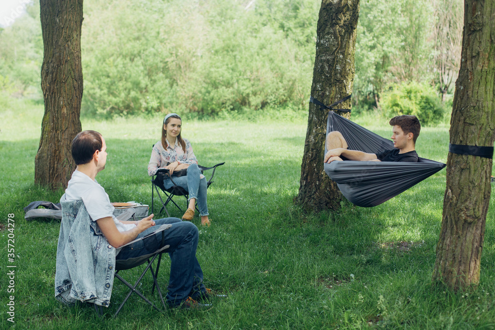 Small group of people enjoying conversation at picnic in accordance with social distancing in summer park. Friends chilling in hammock and on chairs among trees - obrazy, fototapety, plakaty 