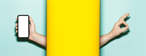 Male hand holding smartphone with white empty mockup on screen. Another hand showing OK gesture, between two studio backgrounds of yellow and aqua menthe colors.