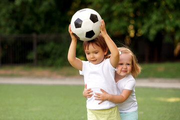 Gym lesson in kindergarten: two toddler children, boy and girl, playing together with soccer ball at football field, girl is holding boy from behind, preventing him from throwing ball. Copy space