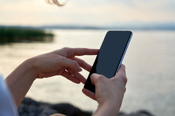 Close-up photograph of a blue screen smartphone held by a woman. In the background the sunset light and the sun reflecting on the blue water of Lake Garda.