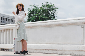 Beautiful traveler asian woman wearing white shirt, long skirt and hat with large luggage on white bridge background in Bangkok city Thailand, travel concept.