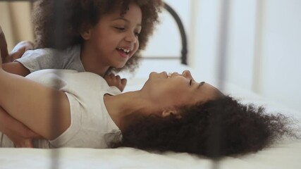 Wall Mural - Cute funny little african american child daughter tickling happy mother playing together lying on bed, cheerful mixed race family small kid girl and black mom embrace cuddle laugh relax in bedroom