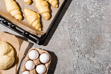 Wall Mural - top view of uncooked croissants on baking tray near eggs and dough on concrete grey surface