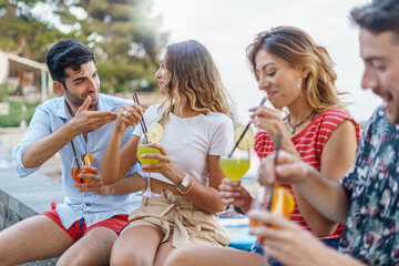 Group of trendy happy young people talking and having fun drinking tropical cocktails together sitting on a bench in the summer