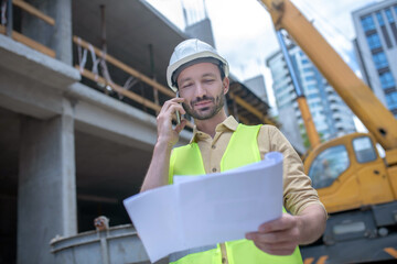 Building worker in helmet and yellow vest holding blueprint, talking on the phone