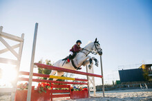 Low Angle View Of Teenage Jockey Girl Riding White Horse Over Hurdle On Training Ground Against Clear Sky