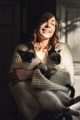 Wall Mural - Attractive girl embracing her Siamese cats. Indoor portrait of cute woman playing with her two pets.