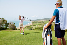 Caddy Watching Woman Tee Off On Golf Course Overlooking Ocean