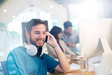 Wall Mural - Smiling creative businessman with headphones talking on cell phone in office