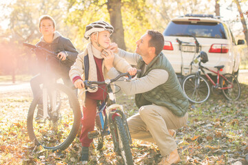 Father fastening helmet of son on bicycle in autumn woods