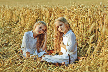 Sitting young twins sisters with book in golden field on a summer sunny day. Portrait of two blonde girls posing on nature background. Summer vacation in countryside.