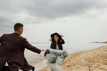 Happy young couple have fun on the beach. Young attractive  happy man and stylish woman in blue dress and black hat running together on beach on vacation, traveling. Romantic couple. Copy space.