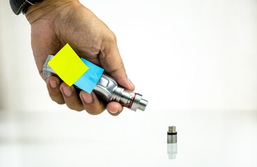 Conceptual of recent uncertainty of safety of e-cigarettes. Isolated close-up of hand holding vape kit battery with atomizer over reflective white besides mouthpiece.  Focus on labels.