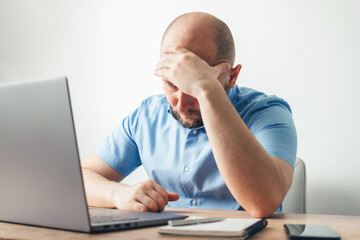 Frustrated and tired young man working online on his laptop at the wooden table in a home office, feels headache, stress and problems. Business, freelance or study concept