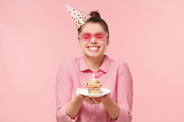 Excited funny teenage girl wearing colored glasses and birthday hat, holding cupcake on plate with two hands, making wish with closed eyes, isolated on pink background