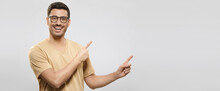 Banner Of Young Smiling Guy In Beige T-shirt And Eyewear, Showing Commercial Offer On Right, Pointing To It With Both Hands, Isolated On Gray Background