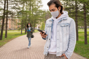 Wall Mural - Modern man in denim jacket and facial mask standing in park and checking phone message