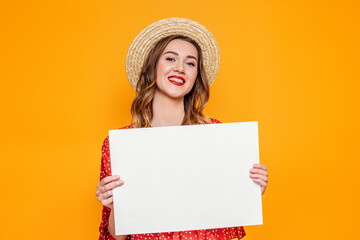 A young woman in a summer red dress, a straw hat holds an A4 empty paper poster and smiles. Happy girl shows a mockup poster, canvas, white sheet of paper isolated over orange background