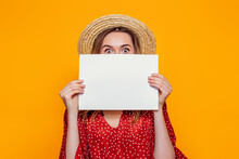 A Young Shocked Girl In A Summer Red Dress, A Straw Hat, Holds An A4 Empty Poster And Covers Her Face. Surprised Woman Shows A Poster, Canvas, White Sheet Of Paper Isolated Over Orange Background