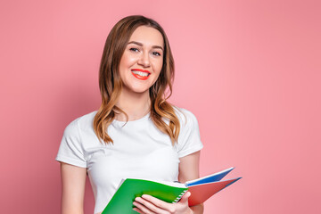 Student girl holding an exercise book, notebook and smiles isolated over pink background, copy space