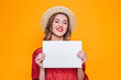 © Evgenia - Portrait of a girl in a red dress, a straw hat with red lips smiling and holding in her hands an A4 format poster isolated over orange background, mockup for greeting card text