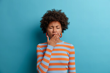 Wall Mural - Tired exhausted Afro American woman feels bad after hard day at work, covers mouth and has sleepy expression, wears casual striped jumper, isolated on blue background. Restless and sleepiness concept