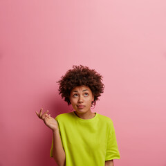 Wall Mural - Isolated shot of ethnic curly woman shrugs shoulders and stands hesitant, raises palm, looks above confused, wears green t shirt, isolated on pink background, empty space for your information