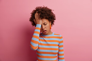 Wall Mural - Portrait of unhealthy young curly woman has severe headache, keeps hand on forehead, feels unwell because of migraine, being stressed and worried, keeps eyes closed, isolated on pink background.