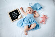 Eleven months old baby girl wearing blue summer dress laying down on white background with letter board and teddy bear.