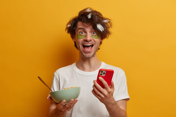 Good morning concept. Cheerful man awakes in good mood, eats cereals for breakfast, uses modern cell phone, checks email box, has messy hairstyle, feather stuck in hair, wears casual white t shirt