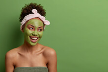 Head Shot Of Smiling Carefree Woman Has Bare Shoulders, Looks Away With Pleasure, Does Skin Care Beauty Facial Routine, Wears Green Mask On Face, Headband, Isolated Over Studio Wall With Blank Space