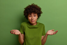 Puzzled Hesitant Young African American Woman Spreads Palms, Feels Unsure, Looks In Bewilderment, Stands Questioned, Wears Green Casual T Shirt In One Color With Background, Takes Hard Decision
