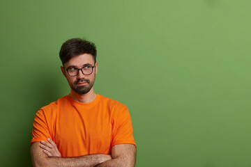 Wall Mural - Thoughtful male model with serious expression, stands with crossed hands and looks away, thinks what to do or makes plans, has thick stubble, isolated on green background, blank space aside.