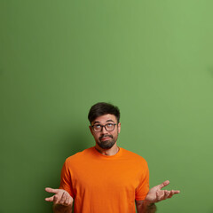 Wall Mural - Vertical shot of questioned clueless man shrugs shoulders and raises palms, has doubtful expression, concentrated above, hesitates what to choose, poses against green wall, empty space upwards