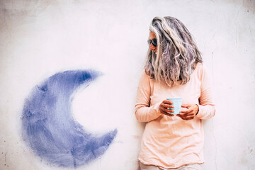 Portrait of adult senior alternative caucasian people woman with long grey and white hair and cup of tea .- white wall wih blue moon in background