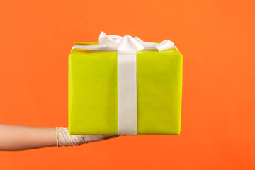 Profile side view closeup of human hand in white surgical gloves holding gift box. sharing, giving or delivery concept. indoor, studio shot, isolated on orange background.