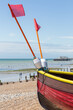 Close-Up View of Bow Section of Small Brightly Coloured Fishing Boat on a Stony Beach.  View of the Ocean in the distance and People on  the Shore. Pier on Horizon