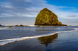 Haystack Rock reflected in the wet sand, with wave foam and blue sky and clouds, Canon Beach, Oregon