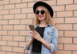 © guas - Portrait of smiling young woman with phone wearing a black round hat, jeans jacket over brick wall background