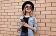 © guas - Portrait of smiling young woman with phone looking away wearing a black round hat, jeans jacket over brick wall background