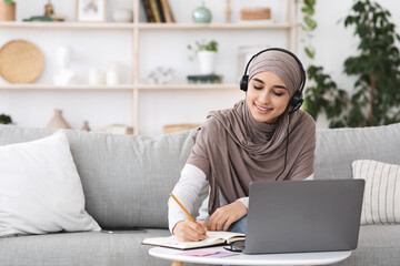 Young arabic woman watching webinar on laptop at home and taking notes